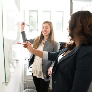 pexels-photo-1181533-1181533 Two professional women discussing ideas on a whiteboard in a modern office setting.
